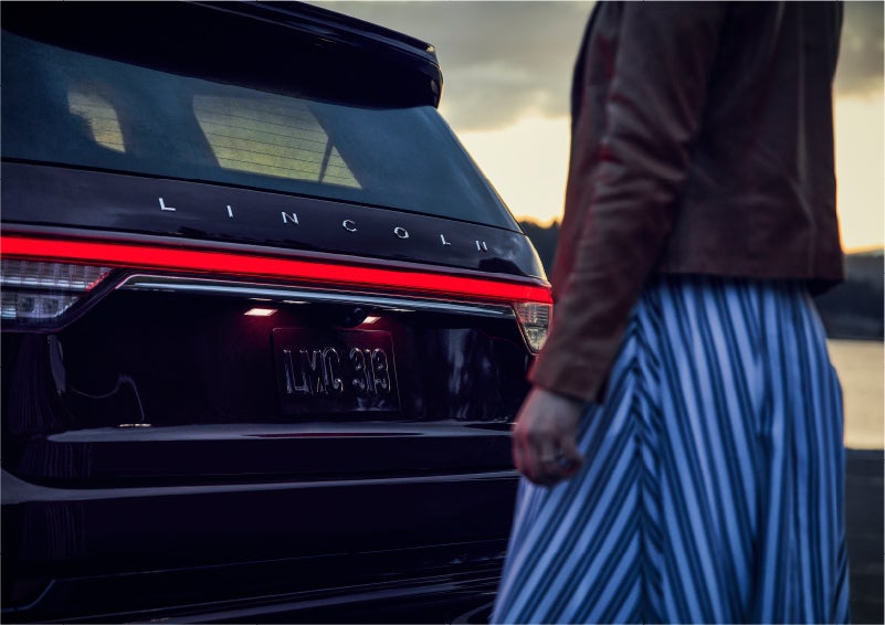 A person is shown near the rear of a 2023 Lincoln Aviator® SUV as the Lincoln Embrace illuminates the rear lights | Astro Lincoln in Pensacola FL